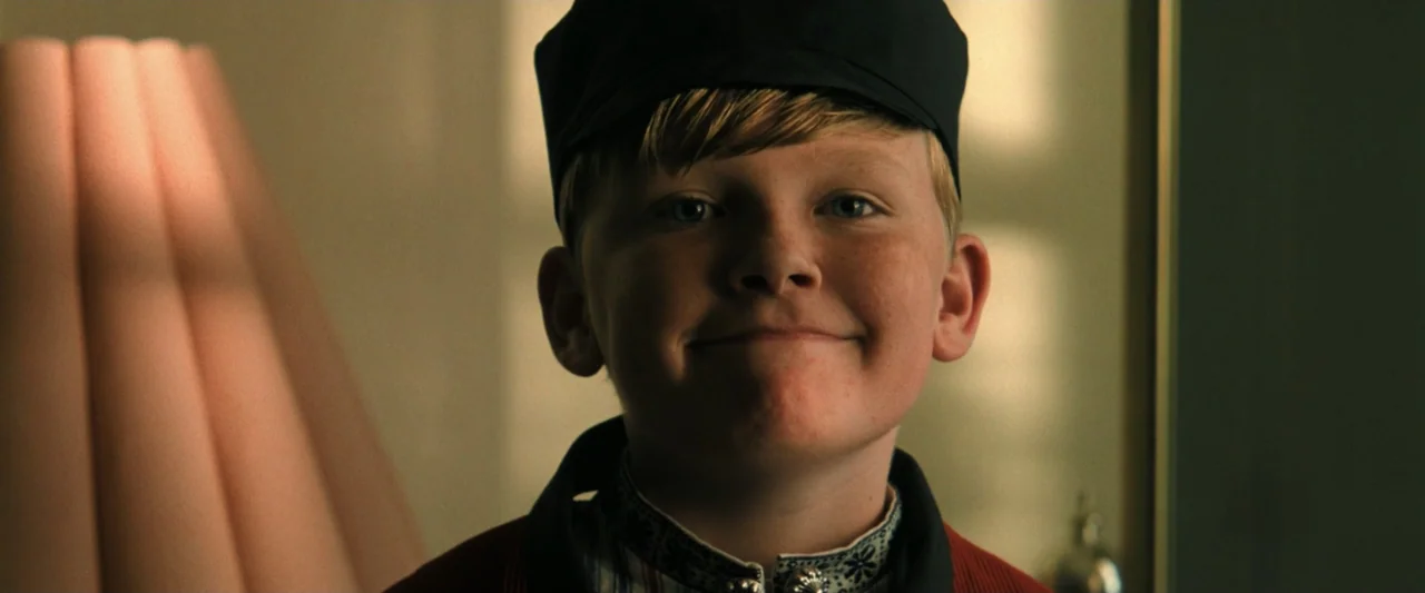 Holland (2025) movie still: closeup — An interior bedroom setting features a child near a lamp with textured wallpaper background. A young boy in a…