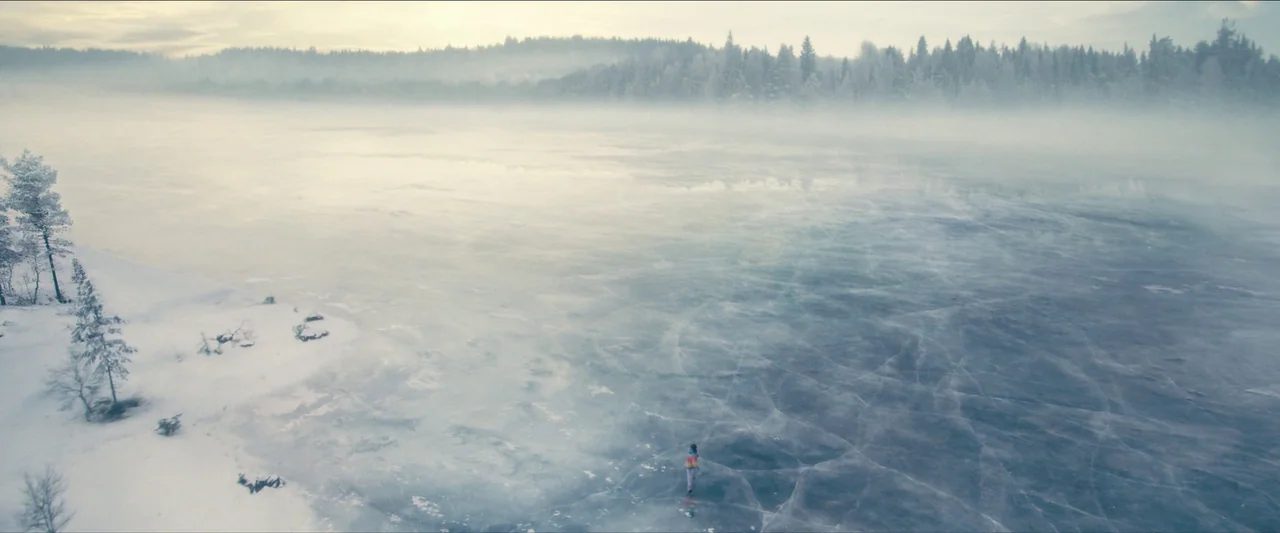 No Time To Die (2021) movie still: wide — A vast, expansive frozen lake covered in thin ice extends toward a dense pine forest. A lone child stands…