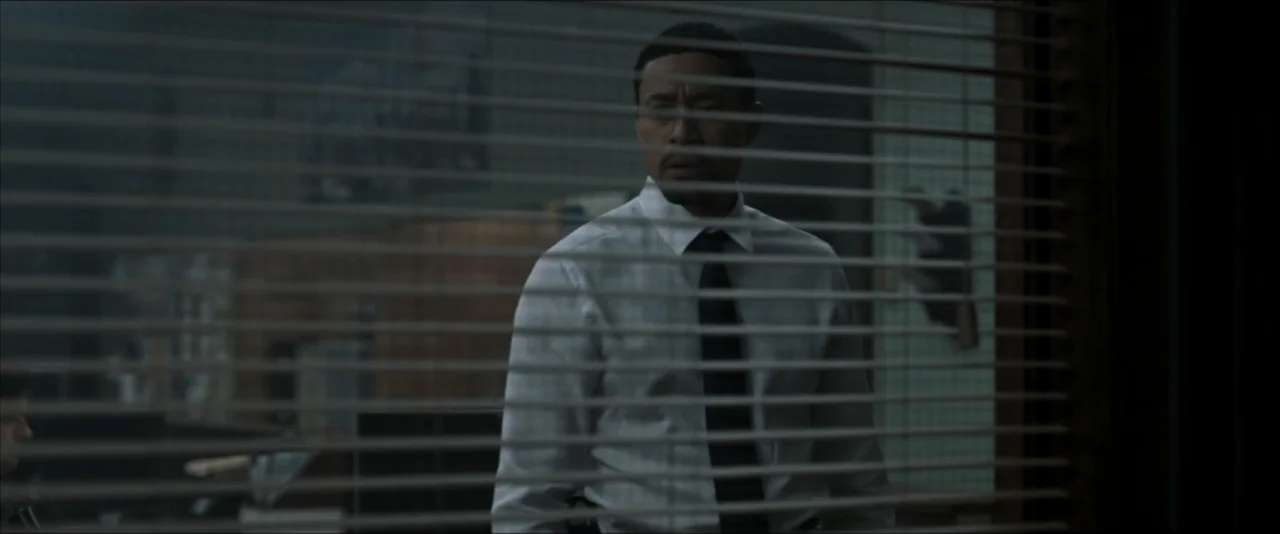 It Feeds (2025) movie still: medium — A man stands behind closed horizontal blinds within a dimly lit office space. He looks through the slats…