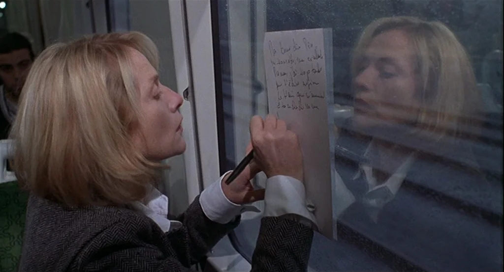 Deux (2002) movie still: medium — A blonde woman sits inside a dimly lit train car at night. She holds a pen to a sheet of paper taped to the dark…