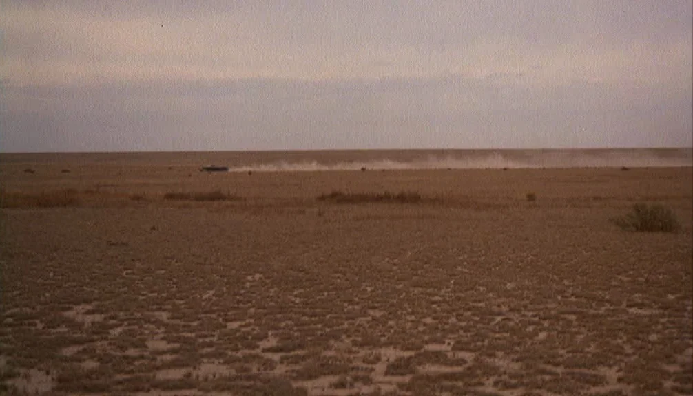 Badlands (1973) movie still: establishing — A vast, barren desert plain stretches to a distant, flat horizon line. A small vehicle moves slowly…
