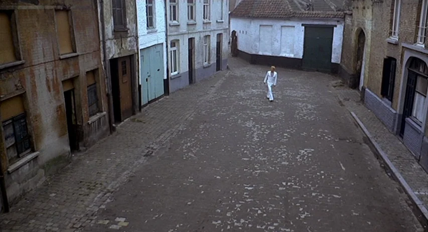 Malpertuis (1971) movie still: wide — A narrow cobblestone street is flanked by old brick and plaster buildings with shuttered windows. A lone man…