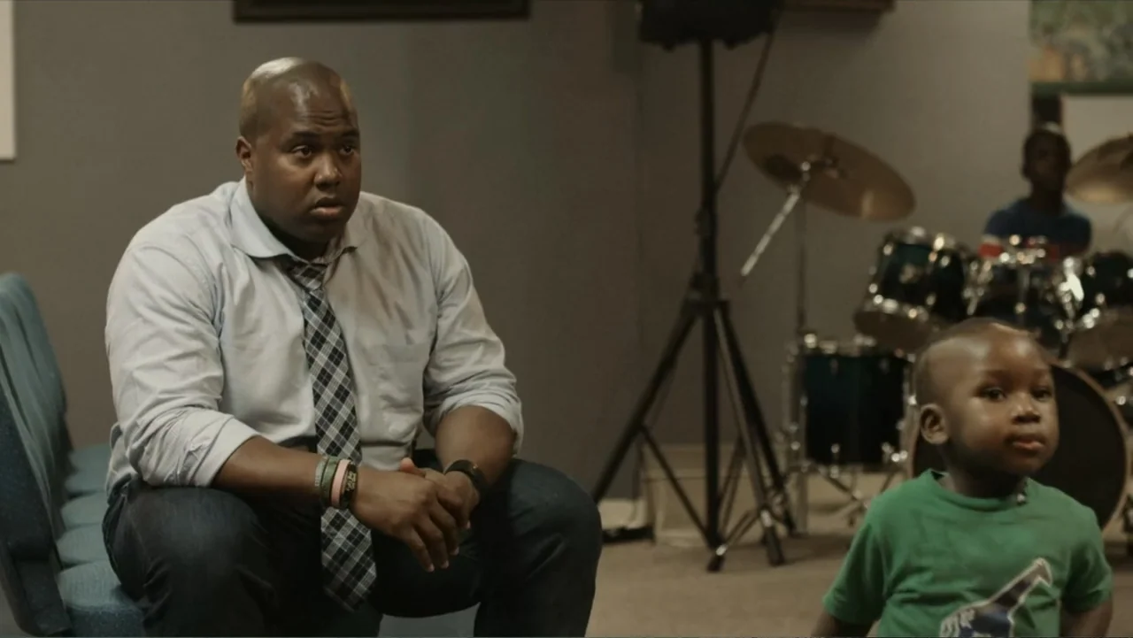Death of a Child (2017) movie still: medium — A dimly lit rehearsal room contains a drum kit and rows of blue chairs. An adult man in a dress shirt…