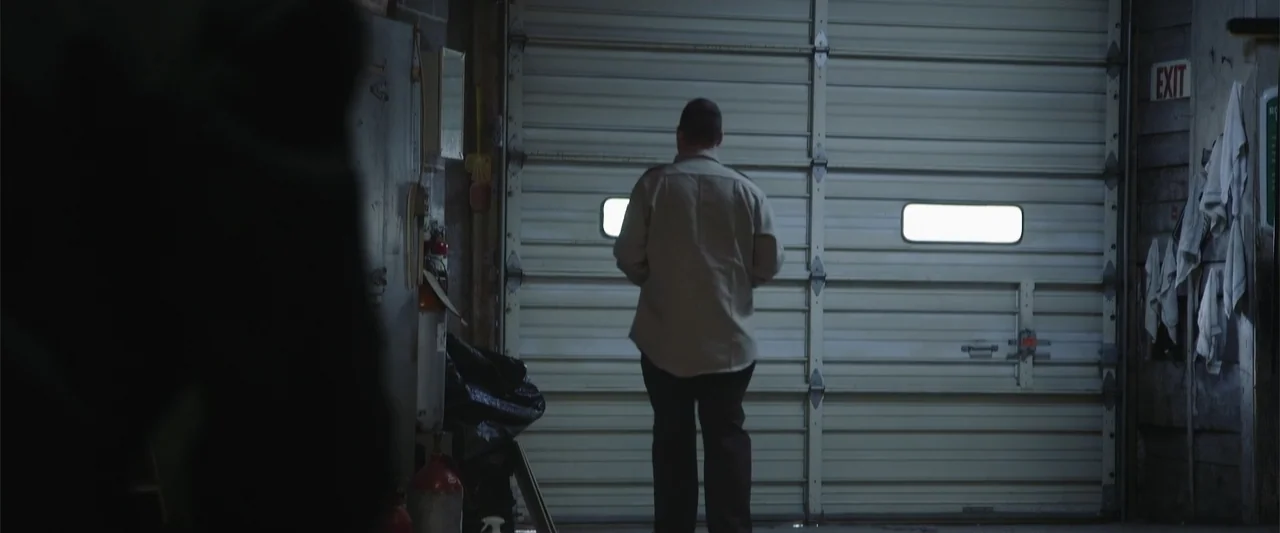 Darkness Waits (2020) movie still: fullbody — A cluttered garage interior with cinder block walls and a metal sectional door. A man stands alone in…