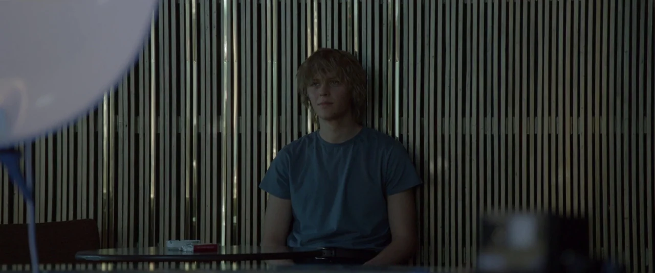 The Lure (2015) movie still: medium — A booth features a textured wood-slat wall paneling behind a seated young man. The man sits at a small…