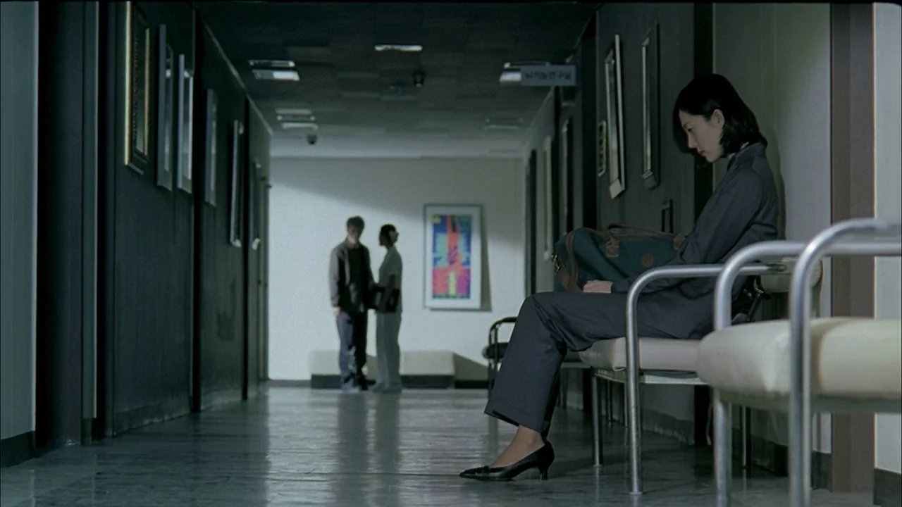 A Tale of Two Sisters (2003) movie still: fullbody — A long, dimly lit hospital corridor features dark walls and framed artwork. A woman sits alone…