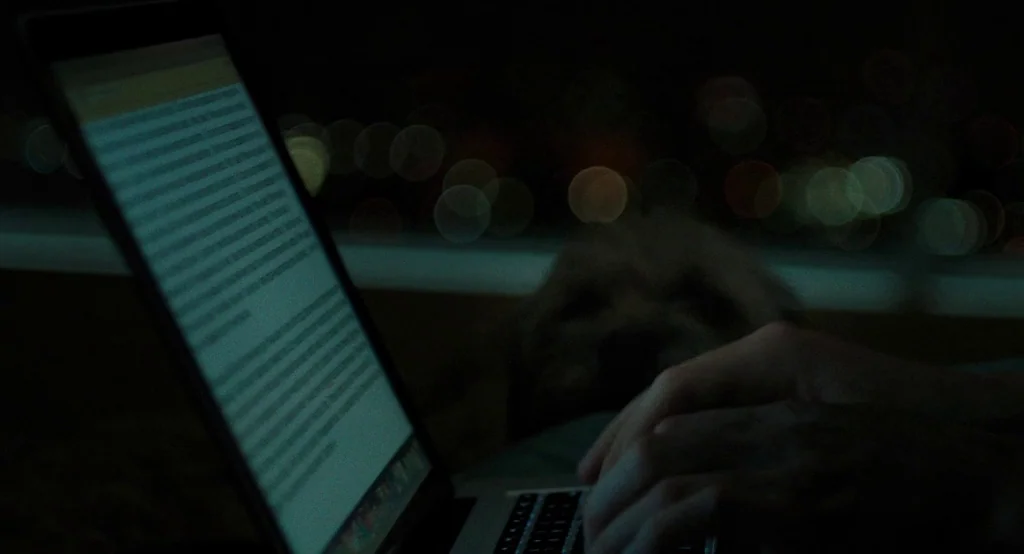Ruby Sparks (2012) movie still: closeup — A dark space at night with a bright computer screen illuminating the foreground. A laptop sits open…