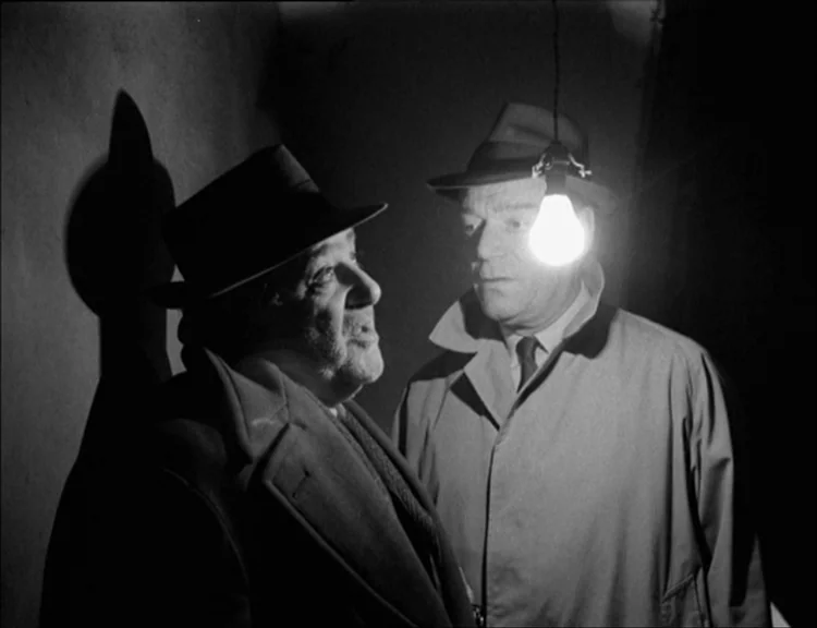 Alphaville (1965) movie still: medium — A narrow, dark interior space with blank walls and a single bare lightbulb. Two men wearing hats and coats…