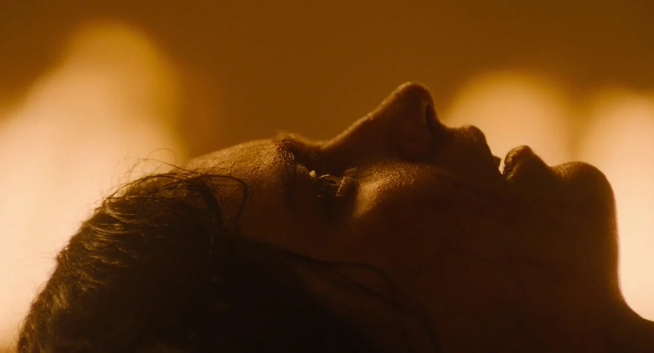 The First Omen (2024) movie still: closeup — A close-up profile view of a woman's face illuminated by intense warm light. The person lies…