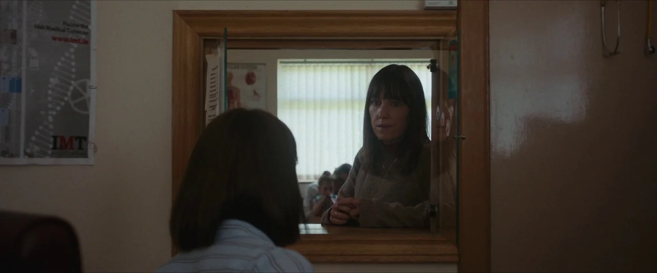 Fréwaka (2024) movie still: medium — A wooden reception window separates a waiting area from an office space. A woman in a brown sweater speaks to a…