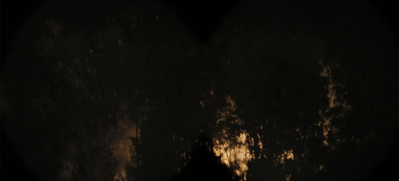 The Tank (2025) movie still: wide — A dark, dense cluster of trees is framed through binoculars with visible reticles. The foliage is lit by…