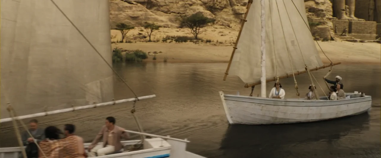 Death on the Nile (2022) movie still: wide — Two small wooden sailboats drift along a desert river beside sandy banks and ruins. People in period…
