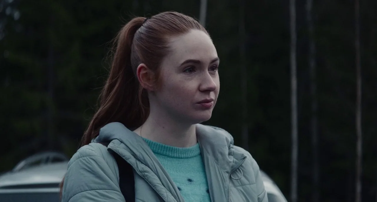Dual (2022) movie still: medium — A woman stands in front of a blurred background of dark, vertical pine trees. The subject looks off-camera with a…