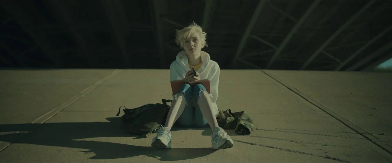 Come True (2020) movie still: medium — A person sits on the smooth concrete ground beneath a massive bridge overpass. The subject holds a small…