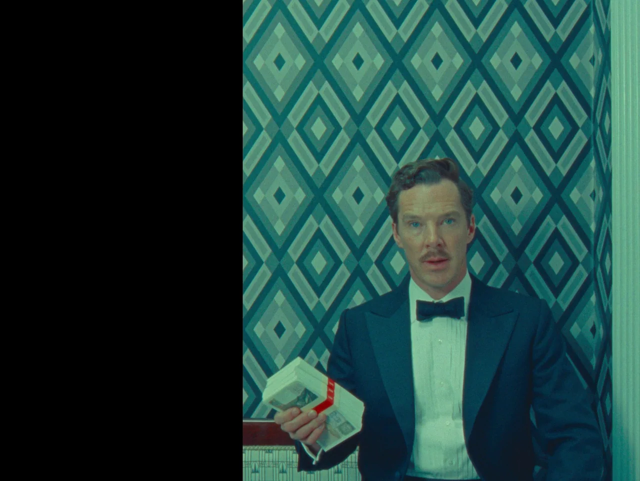 The Wonderful Story of Henry Sugar (2023) movie still: medium — A man stands centered against a patterned geometric wallpaper background. He holds a…