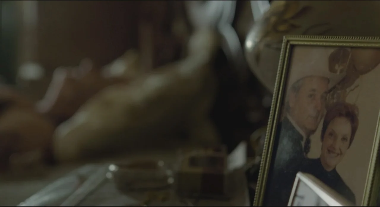 St. Vincent (2014) movie still: closeup — A close-up view shows a framed photograph on a wooden dresser top. The photo depicts a man wearing a…