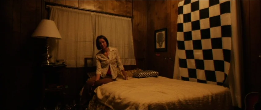 Bubble (2005) movie still: medium — A room features wood-paneled walls and a bed with a large checkered cloth hanging. A woman sits on the edge of…