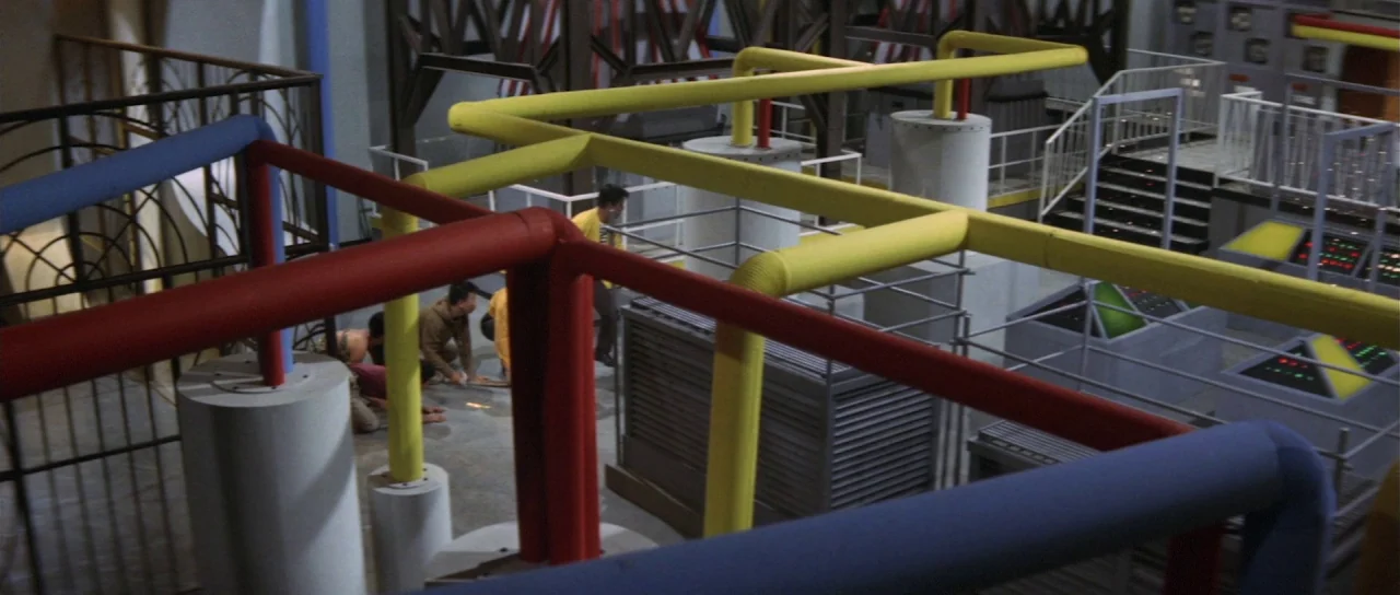 Ebirah Horror of the Deep (1966) movie still: wide — A complex industrial setting features exposed yellow, red, and blue metal pipes overhead. Small…