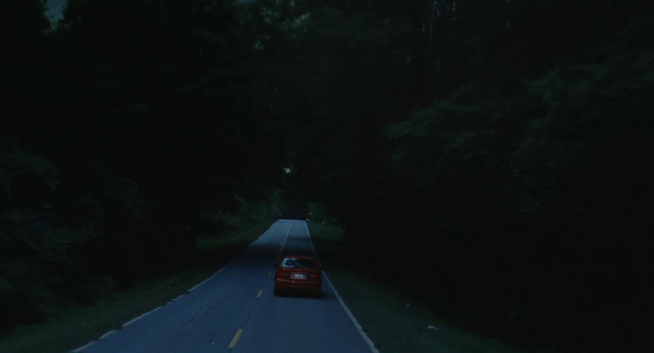 The Giant (2019) movie still: wide — A dark, asphalt road is surrounded by dense, unlit trees on both sides. A single red car drives forward on the…