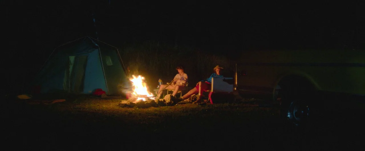 Snack Shack (2024) movie still: wide — A dark outdoor campsite features a tent and a vehicle parked near a fire. Two young men sit in folding chairs…