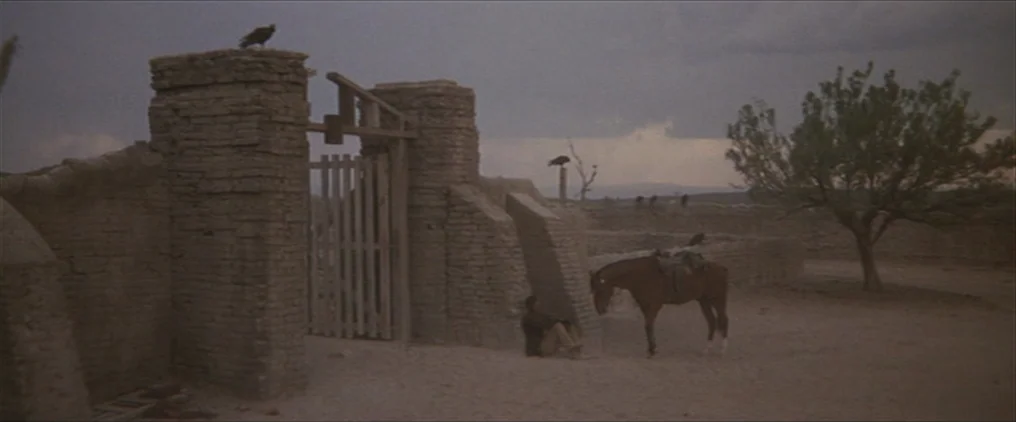 The Wild Bunch (1969) movie still: wide — An adobe structure with stone pillars sits in a desolate, dry outdoor area. A lone man sits against the…