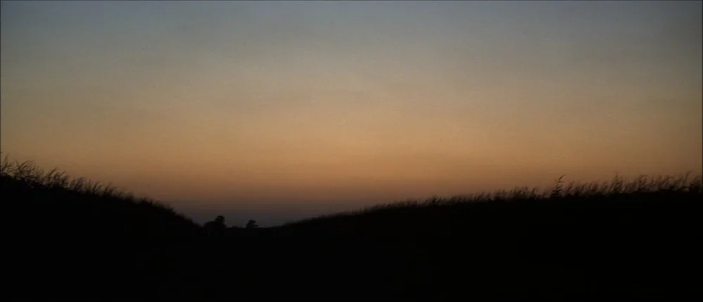 The Straight Story (1999) movie still: establishing — A dark grassy hill stretches across the bottom of the frame against a sunset. The sky displays…
