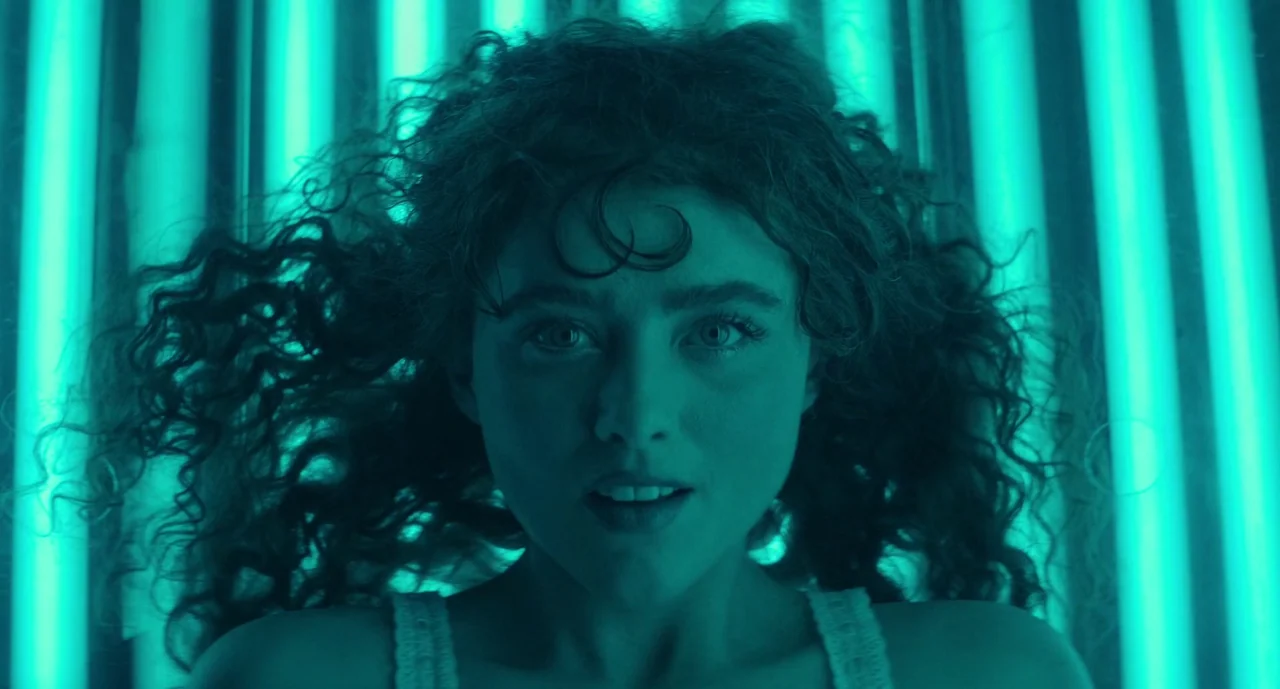 Lisa Frankenstein (2024) movie still: closeup — A close-up of a woman lying inside a tanning bed with illuminated tubes. Her hair fans out around…