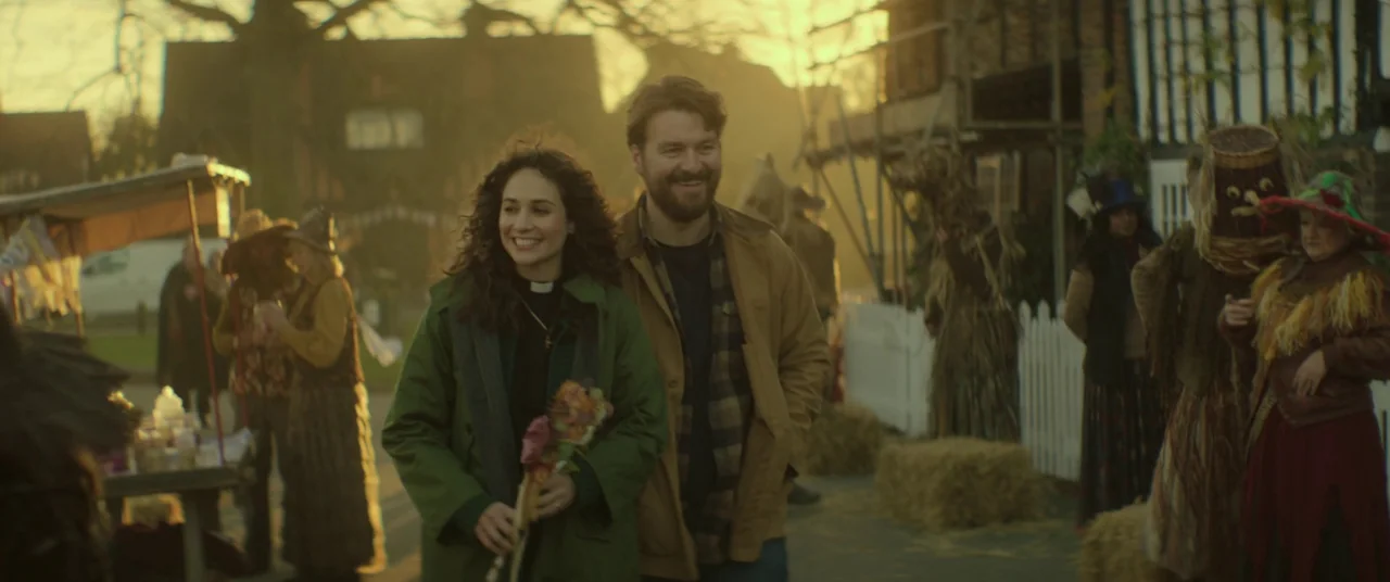 Lord of Misrule (2023) movie still: medium — An outdoor village festival setting features stalls and people in rustic autumnal costumes. A man and…