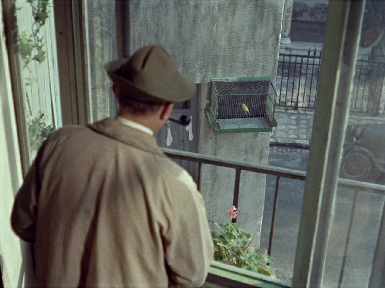 Mon Oncle (1958) movie still: medium — A man stands on a balcony looking at a small birdcage mounted on a wall. The scene includes a view of a…