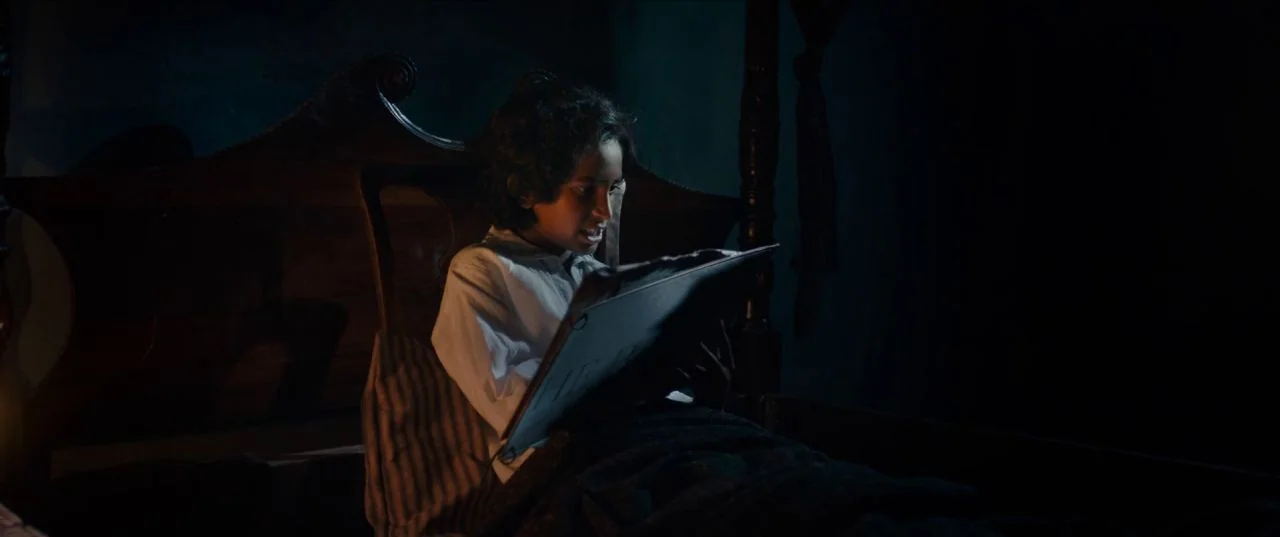 The Personal History of David Copperfield (2019) movie still: medium — A dark, dimly lit bedroom with an ornate wooden headboard visible behind a…