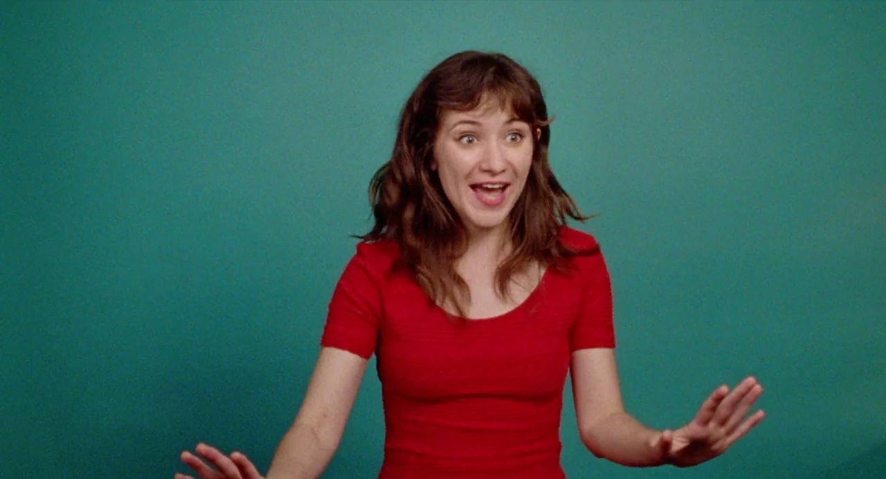 Mr. Roosevelt (2017) movie still: medium — A person stands in front of a flat, solid teal-colored wall in a studio. She holds her hands out in front…