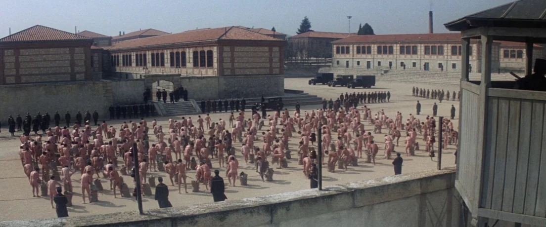 Papillon (1973) movie still: establishing — A sprawling gravel prison yard surrounded by long brick buildings with tiled roofs. A large group of…