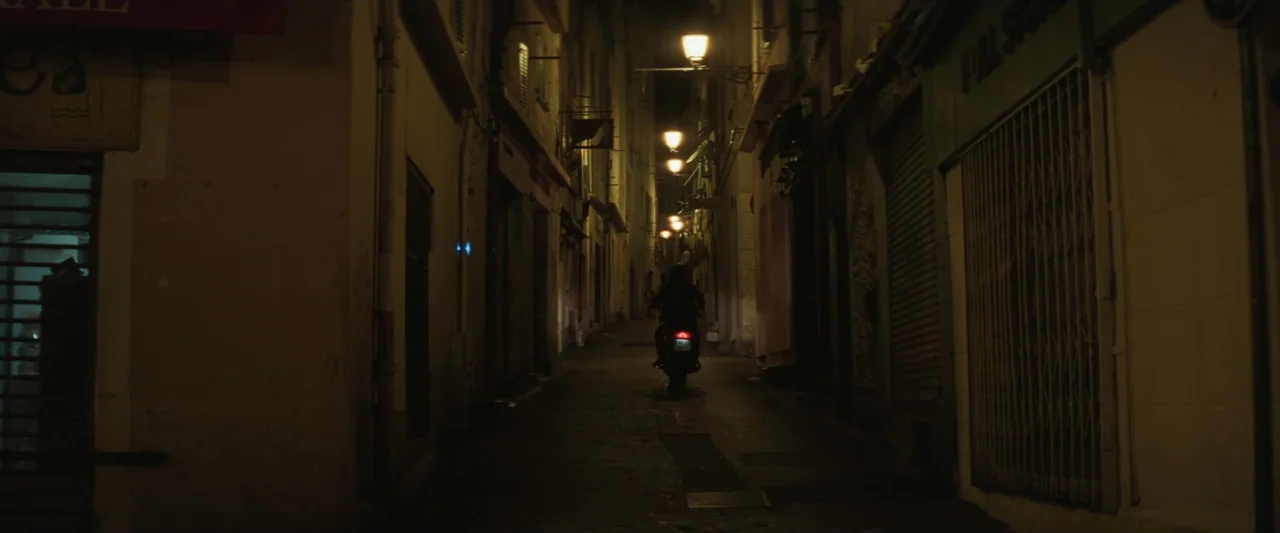 Masquerade (2022) movie still: wide — A narrow stone-walled urban alleyway is illuminated by a row of mounted streetlights. A person on a small…