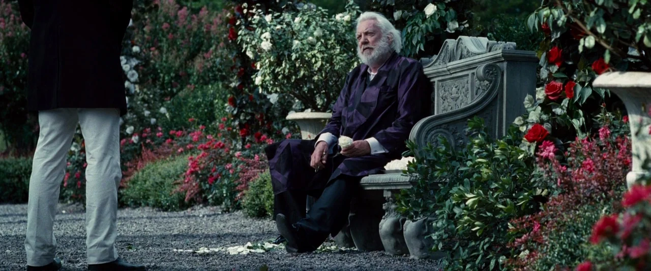 The Hunger Games (2012) movie still: medium — A man sits on an ornate stone bench amidst a dense, formal rose garden. The man holds a single white…