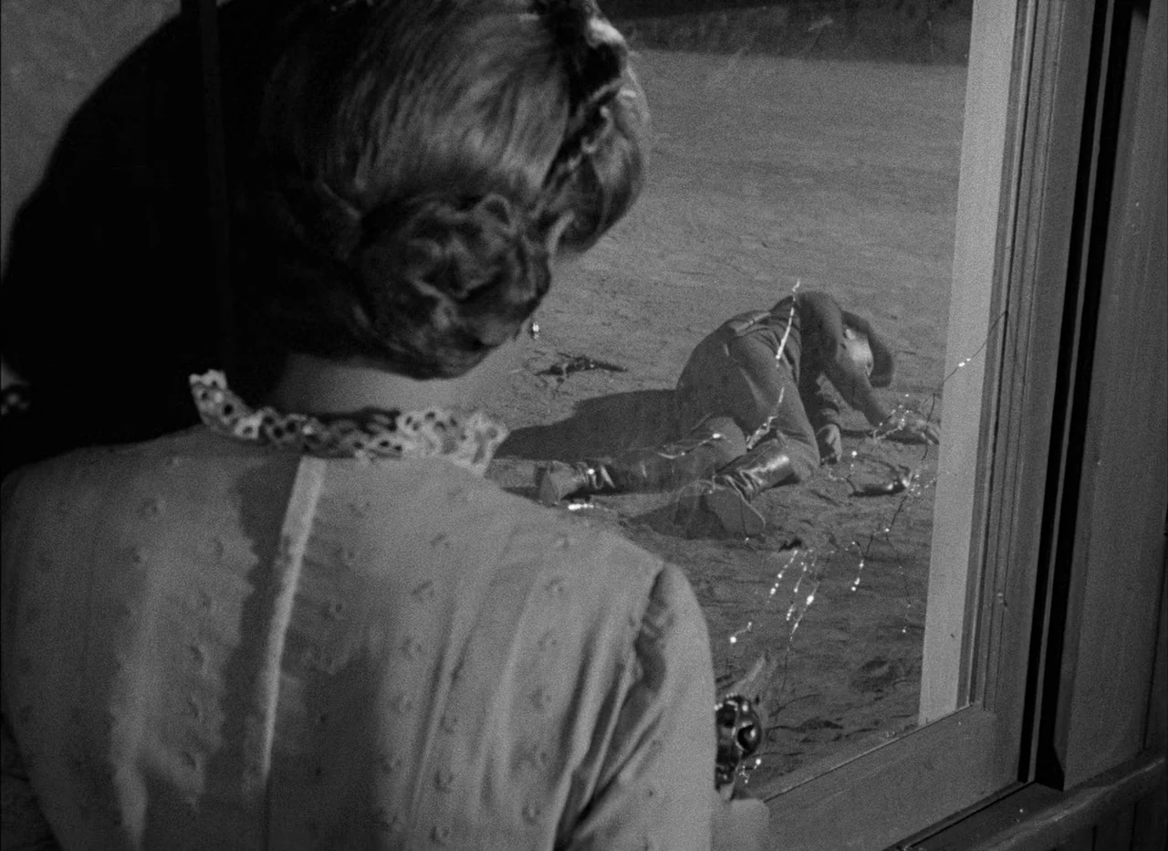 High Noon (1952) movie still: medium — A dark wooden room looks out onto a sun-drenched patch of sand through glass. A man lies motionless on the…