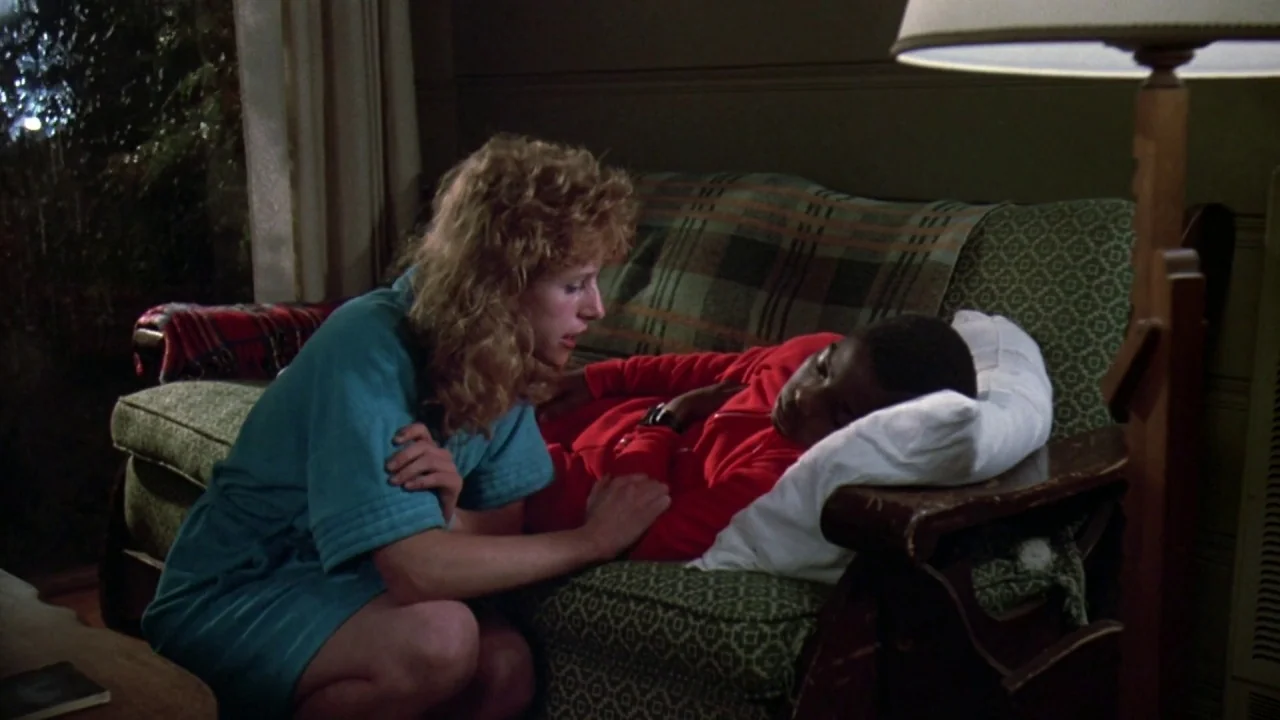 Friday the 13th: A New Beginning (1985) movie still: medium — A dimly lit room features a plaid-covered sofa in front of a window at night. A woman…