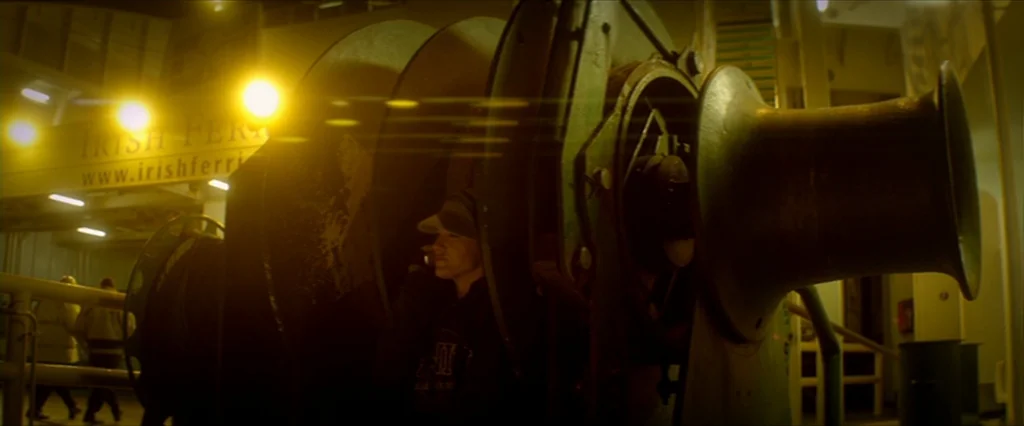 Haywire (2011) movie still: medium — An industrial ferry vehicle deck features large mooring winches and yellow signage overhead. A lone person…