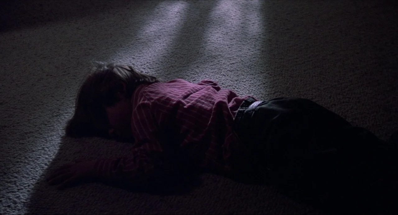 Child’s Play (1988) movie still: medium — A person lies prone on a beige carpet in a dimly lit, shadowed room. A single beam of light illuminates…