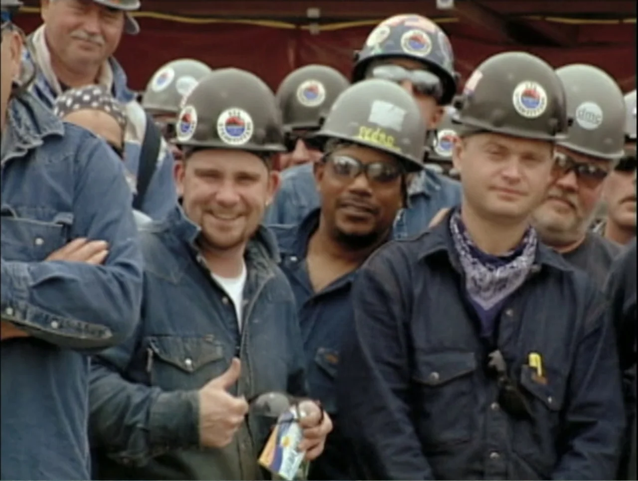 Addiction (2024) movie still: medium — A group of workers stands outdoors at a job site wearing hard hats. The men are grouped together looking…
