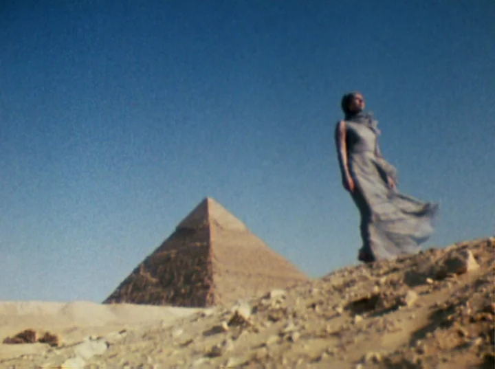 Lucifer Rising (1972) movie still: fullbody — A woman in a flowing dress stands on a sandy hill under a blue sky. The Great Pyramid of Giza rises in…