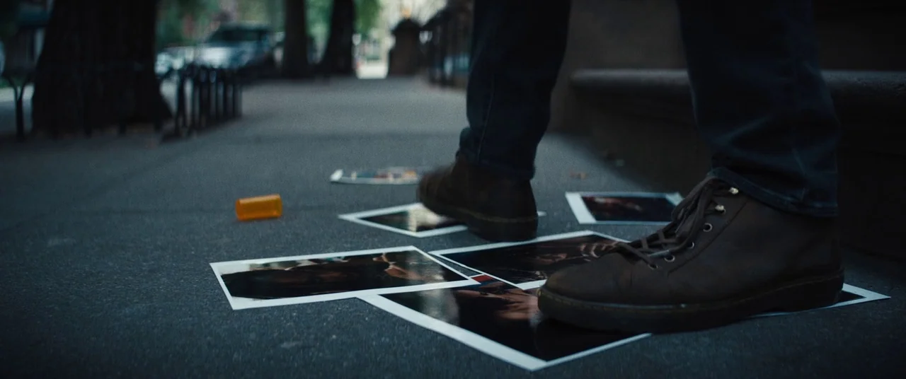 Daniel Isn’t Real movie still: closeup — A dark concrete sidewalk features scattered photographs and a single orange prescription pill bottle. A…