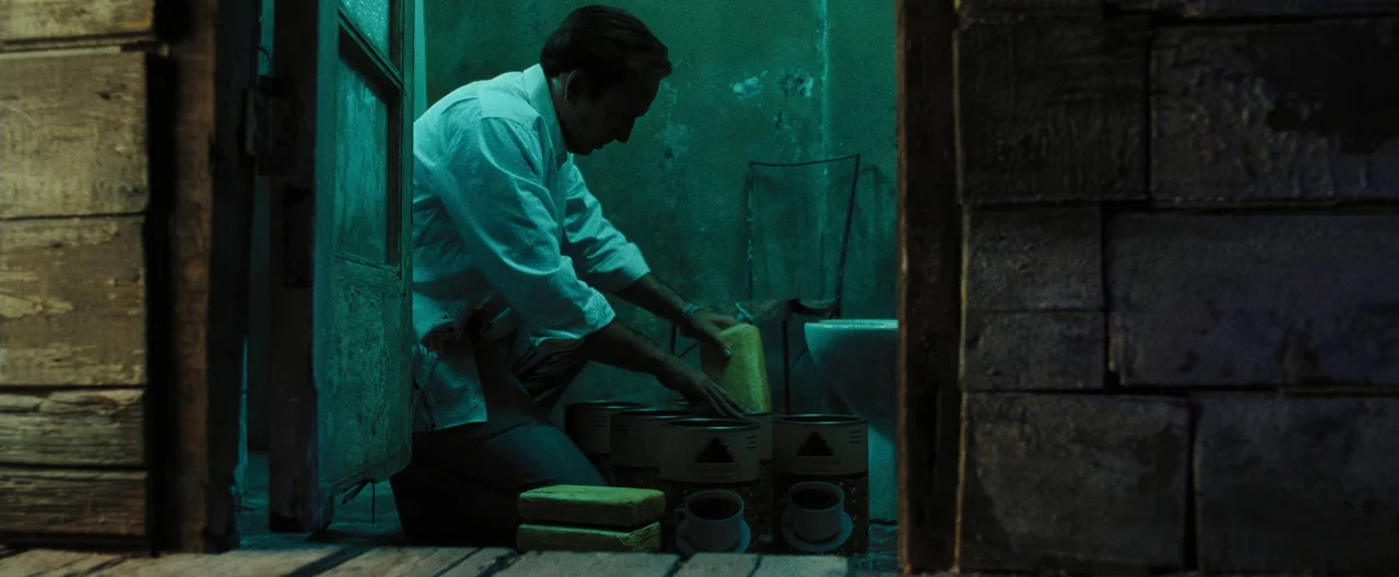 Lord of War (2005) movie still: medium — A dark, cramped wooden room with peeling walls and a visible toilet in back. A man wearing a stained white…