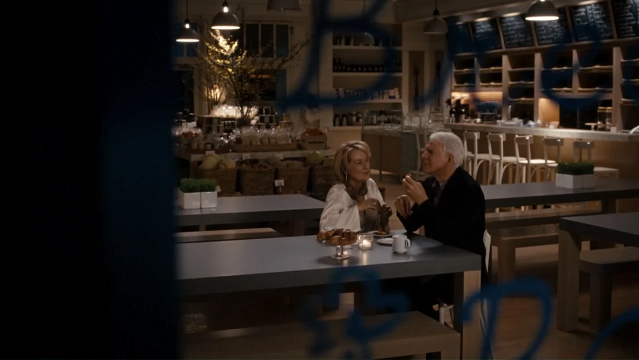 It's Complicated (2009) movie still: medium — A quiet bakery cafe at night features minimalist gray tables and wooden shelving. A man and woman sit…