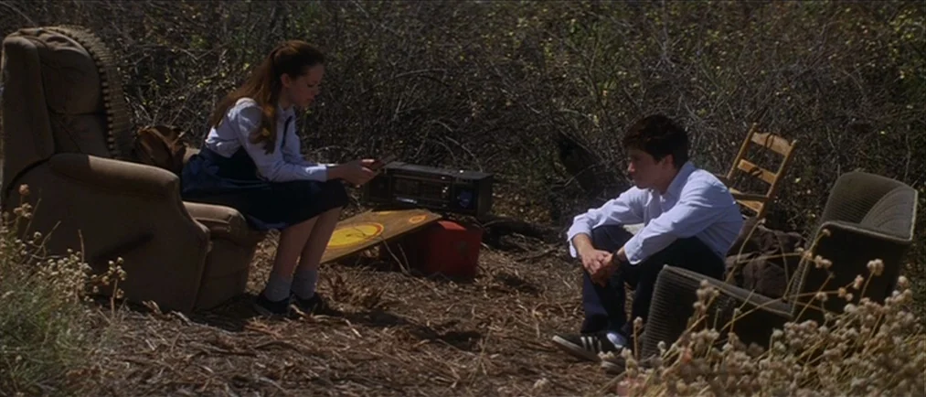 Donnie Darko movie still: fullbody — A pair of teenagers sit on mismatched armchairs in a dry, brush-filled open space. The girl examines a small…
