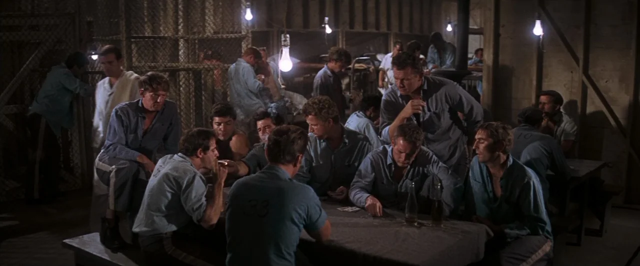 Cool Hand Luke (1967) movie still: wide — A crowded prison barracks interior with rows of beds and a central card table. Several men in uniform blue…