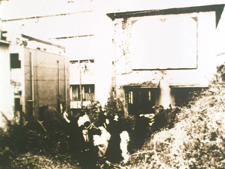 The Reading Machine (1977) movie still: wide — A group of people walks through the rubble of a partially collapsed concrete building. The exterior…