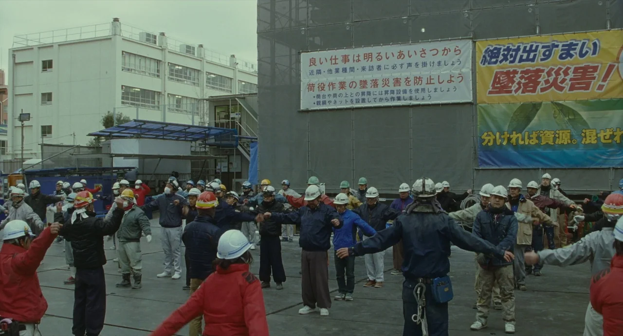Shoplifters (2018) movie still: wide — A large construction site features a crowd of workers in uniform. The workers stand in rows while performing…