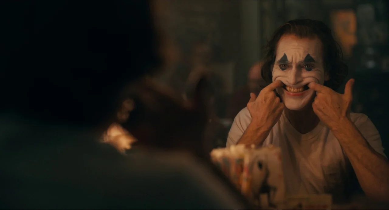 Joker (2019) movie still: medium — An interior space is dimly lit with shadows obscuring the foreground and background. A man applies white face…
