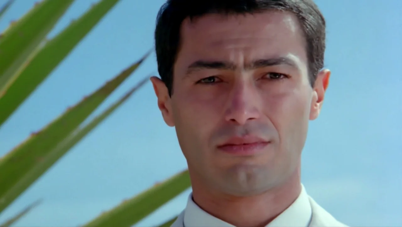 Alexandria…Why? (1979) movie still: closeup — A man stands outdoors under a clear blue sky. A large green palm leaf occupies the left side of the…