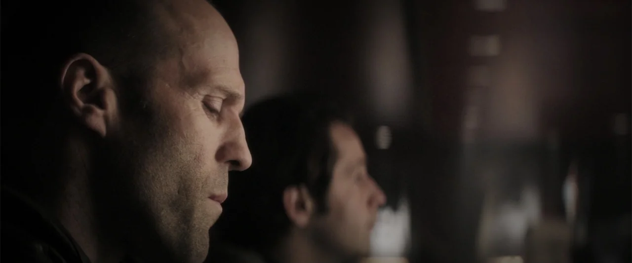 Wild Card (2015) movie still: closeup — A dark, dimly lit bar with out-of-focus background details. Two men sit in profile with their eyes closed in…